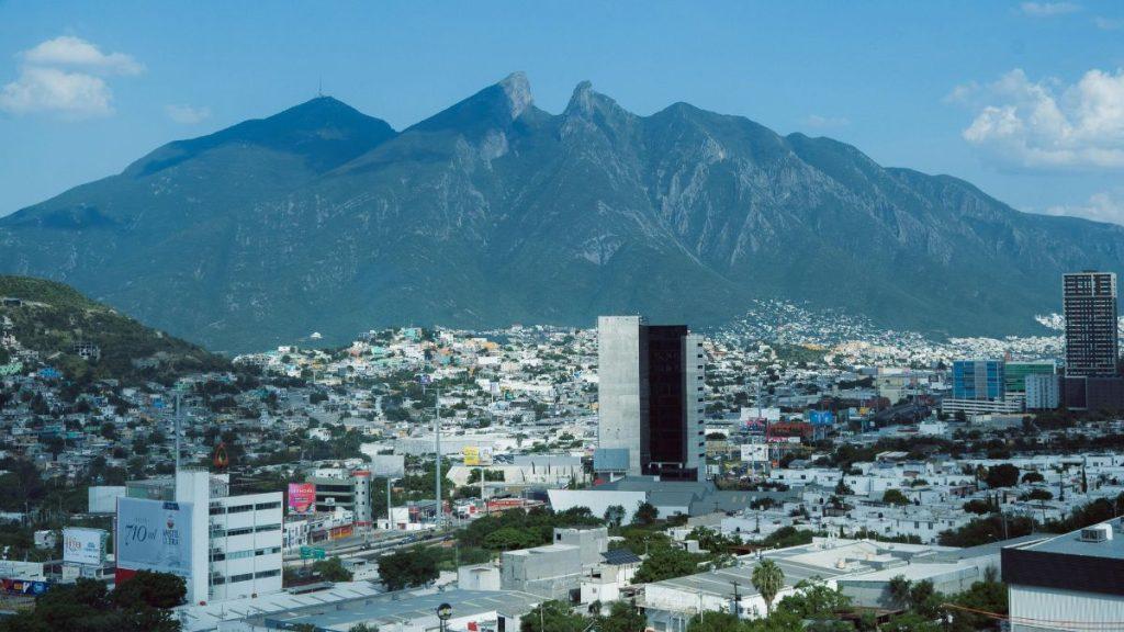 City Skyline with a Tall, Dark Office Building in the Foreground and a Rugged Mountain Range Behind Under a Blue Sky. - Nuevo León Primero