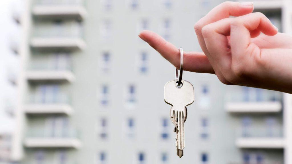 Hand Holding a Keyring with Two House Keys in Front of a Blurred Apartment Building, Suggesting Moving in or Home Access. - Nuevo León Primero