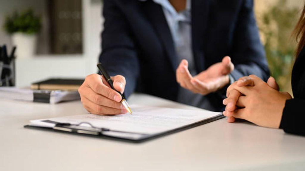 Person Signs a Document with a Pen at a Desk While Another Person Speaks, Indicating a Signing or Agreement Scene. - Nuevo León Primero
