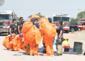 Emergency Responders in Bright Orange Hazmat Suits Gather for a Briefing Near Fire Trucks in a Dusty Training Area. - Nuevo León Primero