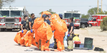 Emergency Responders in Bright Orange Hazmat Suits Gather for a Briefing Near Fire Trucks in a Dusty Training Area. - Nuevo León Primero