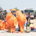 Emergency Responders in Bright Orange Hazmat Suits Gather for a Briefing Near Fire Trucks in a Dusty Training Area. - Nuevo León Primero