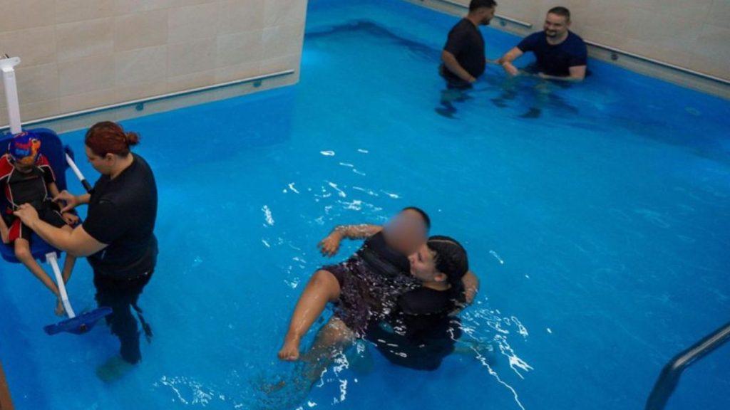 Child in a Flotation Chair Being Assisted by an Instructor at an Indoor Pool; Two Adults Are in the Water Nearby, Engaging in Therapy or Training. - Nuevo León Primero