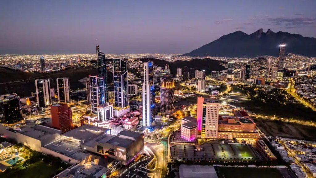 Preventa-quantium-desarrollos-ciudad - Nuevo León Primero Aerial Night View of a Lit City with Tall Skyscrapers, Winding Roads, and a Dark Mountain Range in the Background at Dusk. - Nuevo León Primero