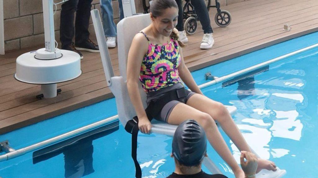 Young Girl in a Bright, Patterned Swimsuit Sits on a Pool Lift Chair at the Poolside, Being Assisted by an Instructor Nearby. - Nuevo León Primero