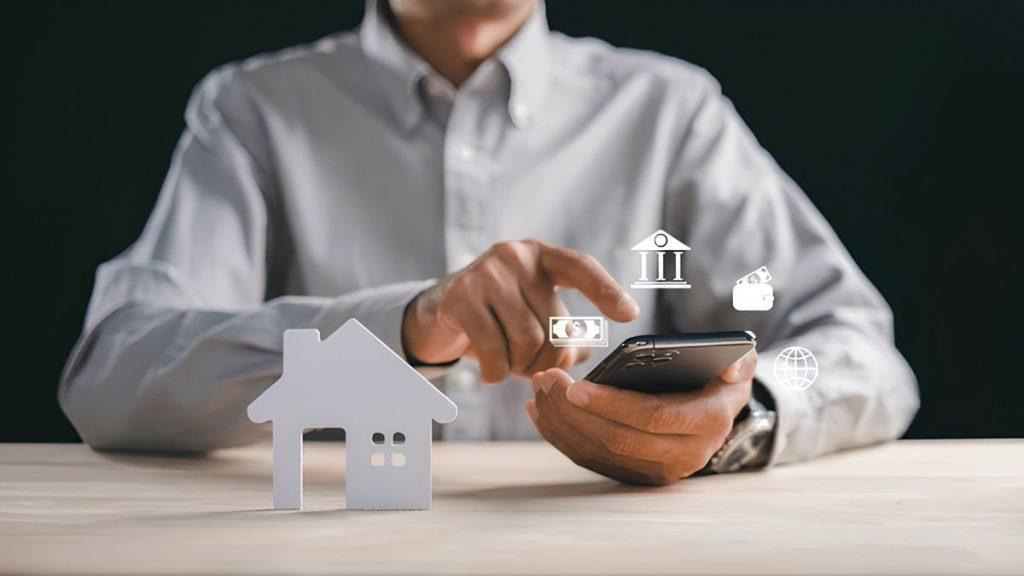 Quantium-desarrollos-cateo-inmobiliaria - Nuevo León Primero Person in a White Shirt Uses a Smartphone While a Small White House Model Sits on the Desk and Digital Finance Icons Hover Around the Screen (bank, Money, Globe). - Nuevo León Primero