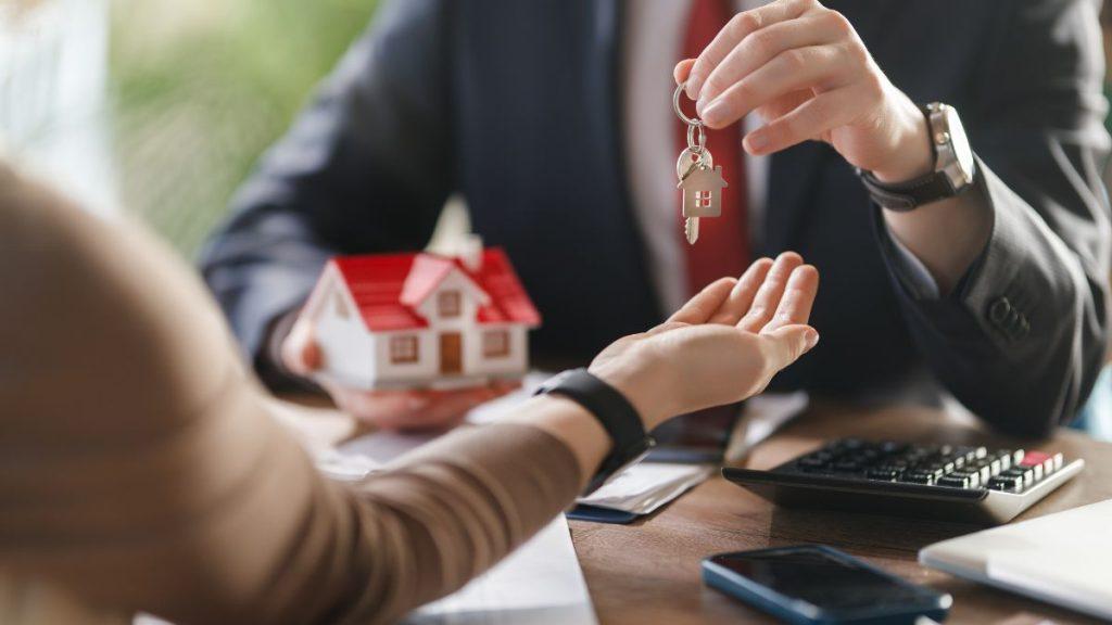 Que-se-necesita-para-comprar-una-vivienda 1 - Nuevo León Primero Real Estate Agent Hands Keys to Client During Home-buying Consultation, with a Small House Model on the Desk and a Calculator Nearby. - Nuevo León Primero