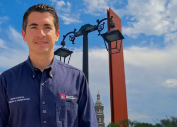 Man in a navy button-down shirt stands outdoors in front of ornate street lamps and a red decorative pillar, blue sky behind him.