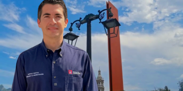 Man in a Navy Button-down Shirt Stands Outdoors in Front of Ornate Street Lamps and a Red Decorative Pillar, Blue Sky Behind Him. - Nuevo León Primero