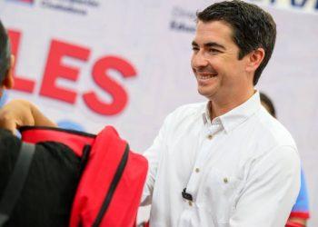 Smiling Man in a White Shirt Hands a Red Backpack to a Person at a Public Event with Large Red Letters in the Background. - Nuevo León Primero