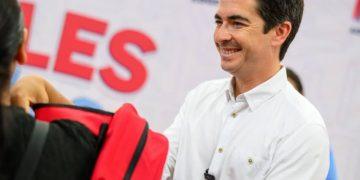Smiling Man in a White Shirt Hands a Red Backpack to a Person at a Public Event with Large Red Letters in the Background. - Nuevo León Primero