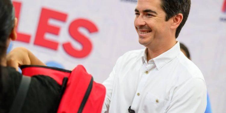 Marcelo Segovia En La Entrega De útiles útiles - Nuevo León Primero Smiling Man in a White Shirt Hands a Red Backpack to a Person at a Public Event with Large Red Letters in the Background. - Nuevo León Primero