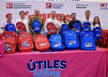 Group of eight adults standing behind a table with red and blue MTY backpacks at a back-to-school event. The banner shows education ministry logos in Spanish context.