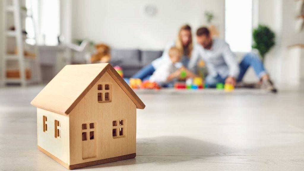 Wooden Toy House on the Floor with a Blurred Background of a Family Playing with Building Blocks in a Living Room. - Nuevo León Primero
