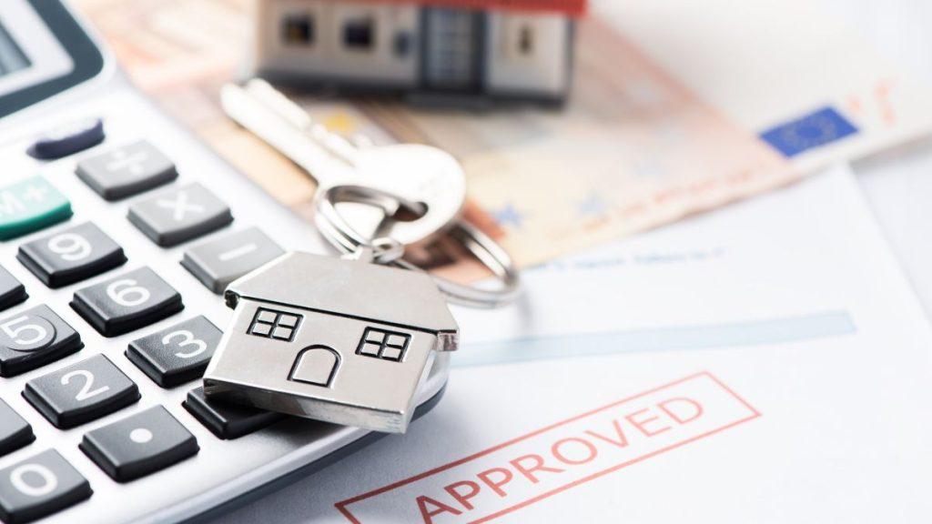 House-shaped Keychain Resting on a Calculator Beside Banknotes, with an “approved” Stamp on a Document, Suggesting Mortgage Approval. - Nuevo León Primero