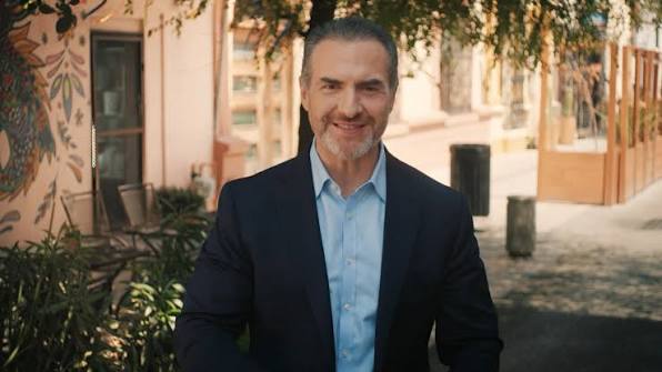 Images - Nuevo León Primero Smiling Man in a Navy Blazer and Light Blue Shirt Standing Outdoors on a Sunlit Sidewalk with Greenery Behind Him. - Nuevo León Primero