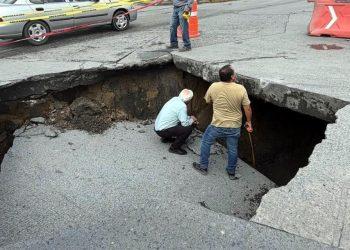 Large sinkhole in a paved road with two workers inspecting the edge, caution tape, and traffic cones nearby.