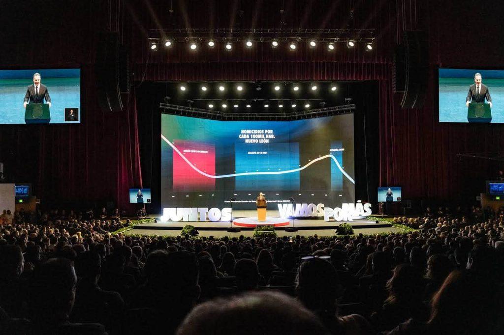 Speaker at Podium on a Large Stage with a Curved Screen Showing a Chart and the Words Juntos Vamos MÁs in Front of a Crowded Auditorium. - Nuevo León Primero