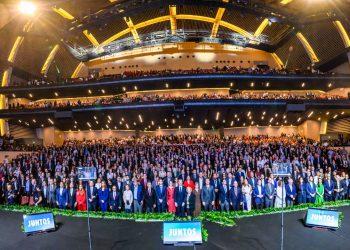 Huge group of people in formal attire posing on a multi-tiered stage in a modern auditorium; 'JUNTOS' signs visible at front.