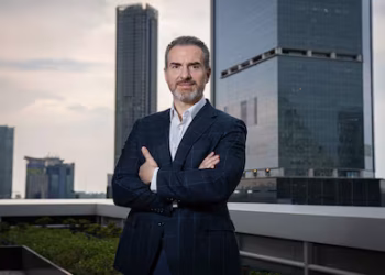 Confident businessman in a dark suit stands with arms crossed on a rooftop, with a city skyline in the background.