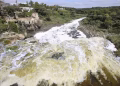 Wide River with White-foamed Rapids Running Between Rocky Banks; on the Left, Old Stone Buildings Cling to a Hillside Amid Green Trees, Under a Blue Sky. - Nuevo León Primero