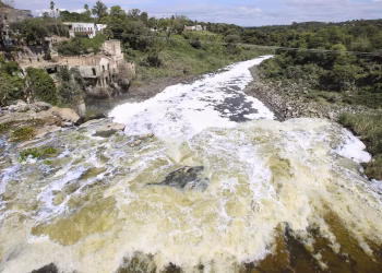 Wide river with white-foamed rapids running between rocky banks; on the left, old stone buildings cling to a hillside amid green trees, under a blue sky.