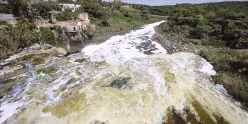 Wide River with White-foamed Rapids Running Between Rocky Banks; on the Left, Old Stone Buildings Cling to a Hillside Amid Green Trees, Under a Blue Sky. - Nuevo León Primero