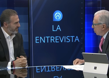 Two Men in Suits Sit Across from Each Other at a Tv Interview Set, with a Blue Backdrop and a Screen Reading 'la Entrevista'. Their Tablet Device Sits on the Desk Nearby. - Nuevo León Primero