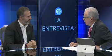 Two Men in Suits Sit Across from Each Other at a Tv Interview Set, with a Blue Backdrop and a Screen Reading 'la Entrevista'. Their Tablet Device Sits on the Desk Nearby. - Nuevo León Primero