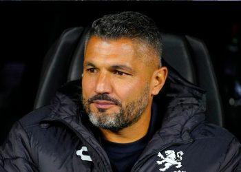 Man with Short Dark Hair and Gray Beard Sits in a Dark Chair, Wearing a Black Quilted Jacket with a White Crest on the Chest. - Nuevo León Primero