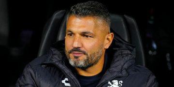 Man with Short Dark Hair and Gray Beard Sits in a Dark Chair, Wearing a Black Quilted Jacket with a White Crest on the Chest. - Nuevo León Primero