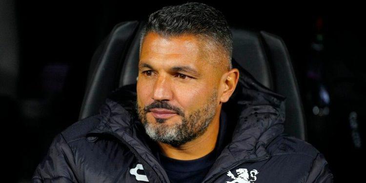 Man with Short Dark Hair and Gray Beard Sits in a Dark Chair, Wearing a Black Quilted Jacket with a White Crest on the Chest. - Nuevo León Primero